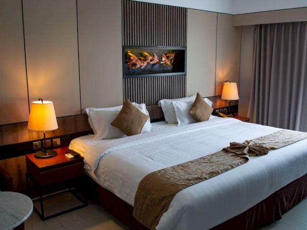 The image shows a neatly made hotel room bed with pillows and a folded towel, surrounded by lamps on bedside tables and a window with curtains.