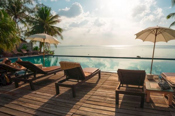 Tropical scene with lounge chairs, parasols, and a serene infinity pool overlooking the ocean under a bright sky. Relaxation paradise.