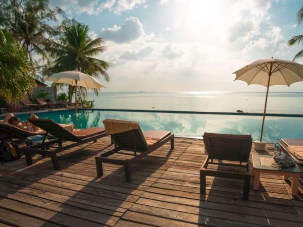 Tropical scene with lounge chairs, parasols, and a serene infinity pool overlooking the ocean under a bright sky. Relaxation paradise.