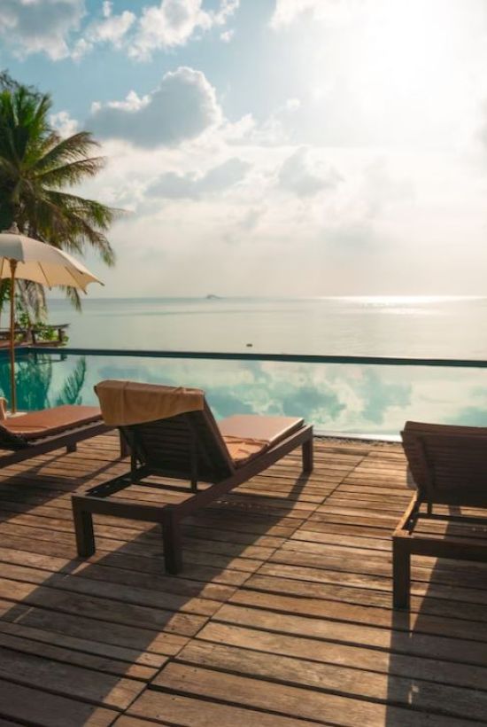 A serene poolside scene with lounge chairs, umbrellas, and palm trees overlooking a calm body of water, under a partly cloudy sky at sunrise or sunset.