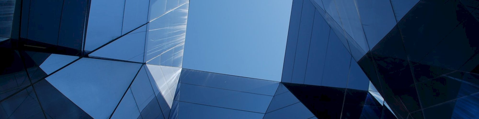 The image shows a view looking up at a blue sky through modern, angular, reflective glass skyscrapers creating an artistic geometric pattern.
