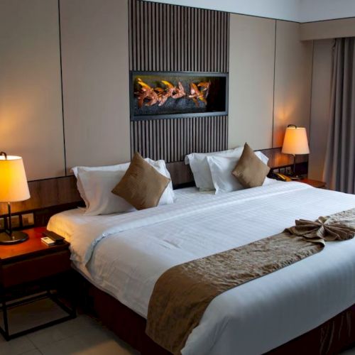 A cozy hotel room with a large neatly made bed adorned with pillows, bedside tables with lamps, wall decor, and a window with curtains.