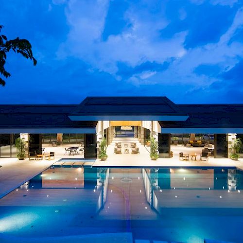 A luxurious resort featuring an expansive pool and poolside seating at dusk, lit with ambient lighting, and surrounded by greenery and open architecture.