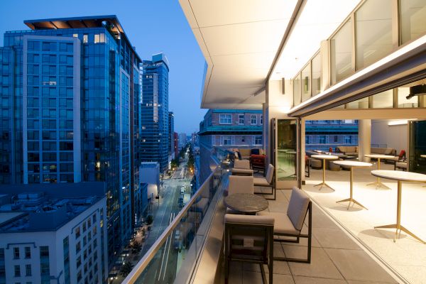 A modern cityscape at dusk, viewed from a high-rise balcony with outdoor seating and tables, overlooking surrounding buildings and a street.