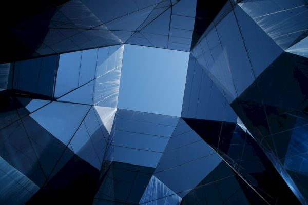 The image depicts an upward view of modern, reflective glass buildings arranged to form a geometric pattern with a clear blue sky at the center.