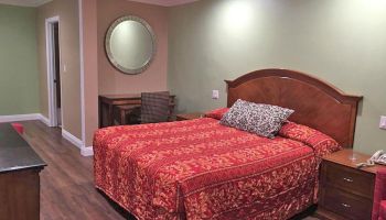 A bedroom with a double bed covered in red bedding, a nightstand, wall mirror, small desk with chair, wooden floor, and green walls.