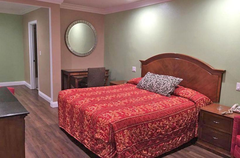 A bedroom with a double bed covered in red bedding, a nightstand, wall mirror, small desk with chair, wooden floor, and green walls.