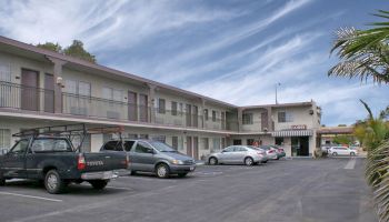 This image shows a two-story motel with a parking lot in front, where multiple vehicles are parked. The sky is blue with scattered clouds.