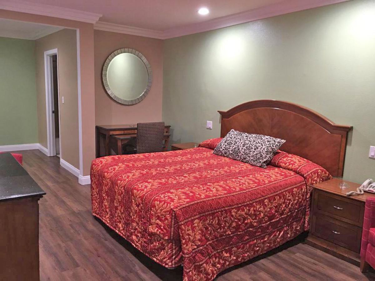 A bedroom with a large bed featuring a red patterned bedspread, a round mirror, a small desk with a chair, and a nightstand.