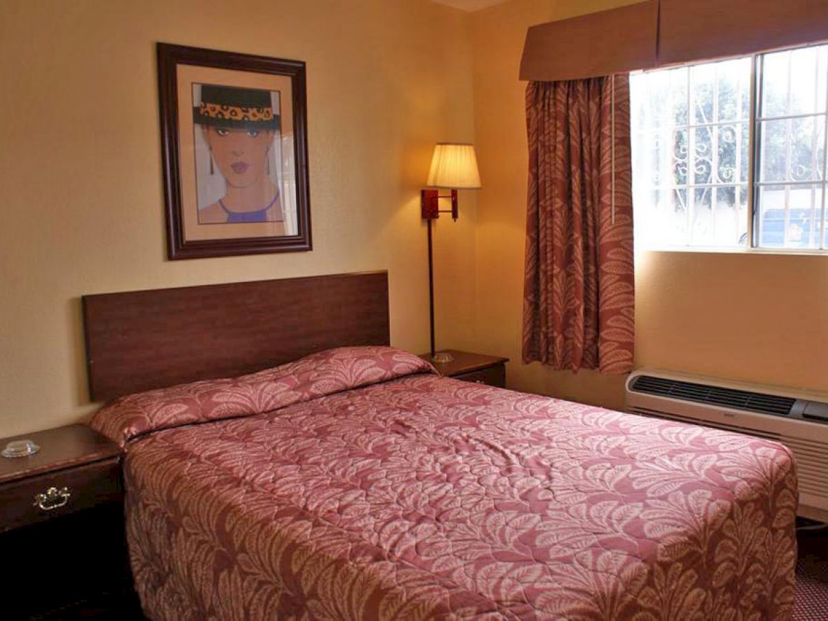 A small, simple bedroom with a double bed, floral bedspread, framed artwork above, side tables, a lamp, curtains, and a window with light streaming in.