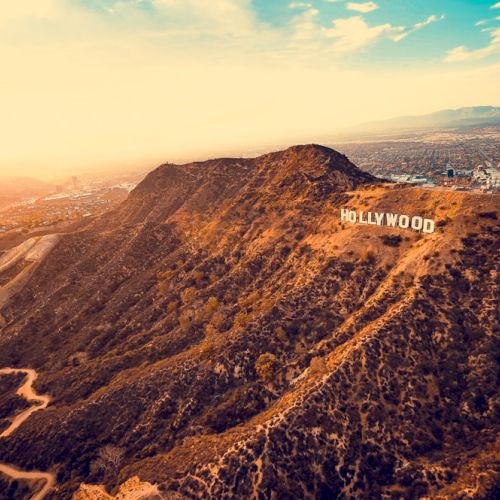 The image shows an aerial view of the iconic Hollywood sign on a hillside with a cityscape and a colorful sky in the background, taken at sunset.