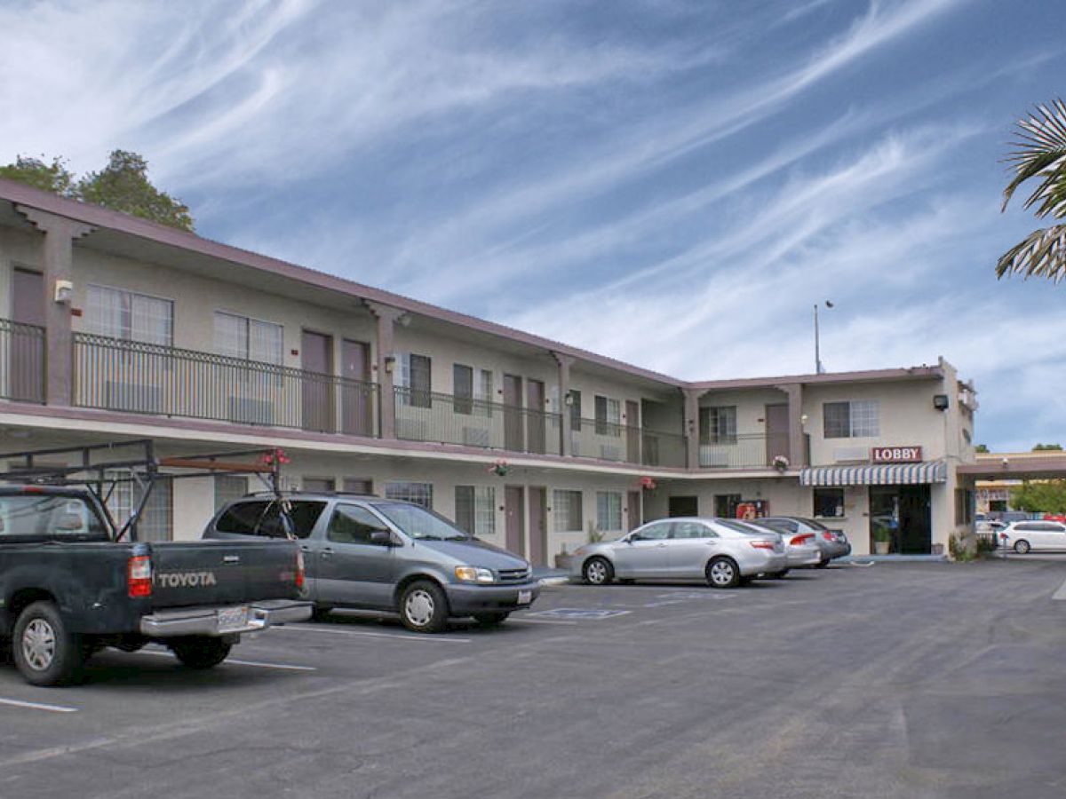 The image shows a parking lot in front of a two-story hotel or motel with several vehicles parked. The sky is partly cloudy, creating a serene ambiance.