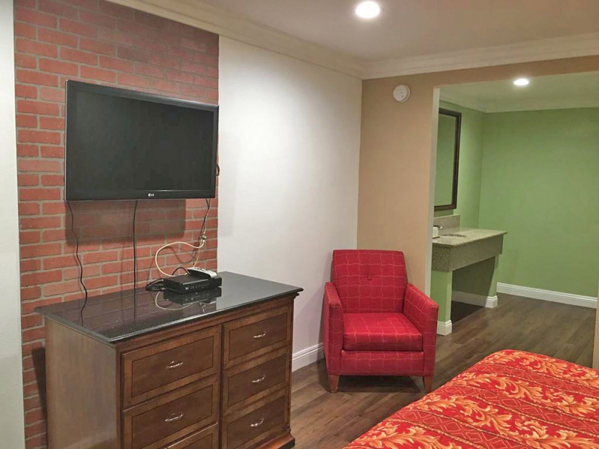 The image shows a hotel room with a wall-mounted TV, a wooden dresser, a red armchair, and a partial view of a bathroom vanity area.