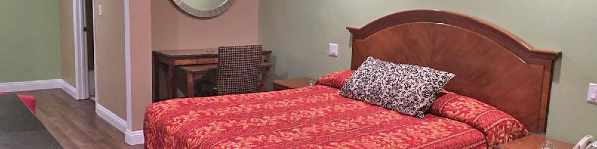 The image shows a bedroom with a red-patterned bedspread, wooden headboard, nightstands, mirror, dresser, and a chair at a small desk.