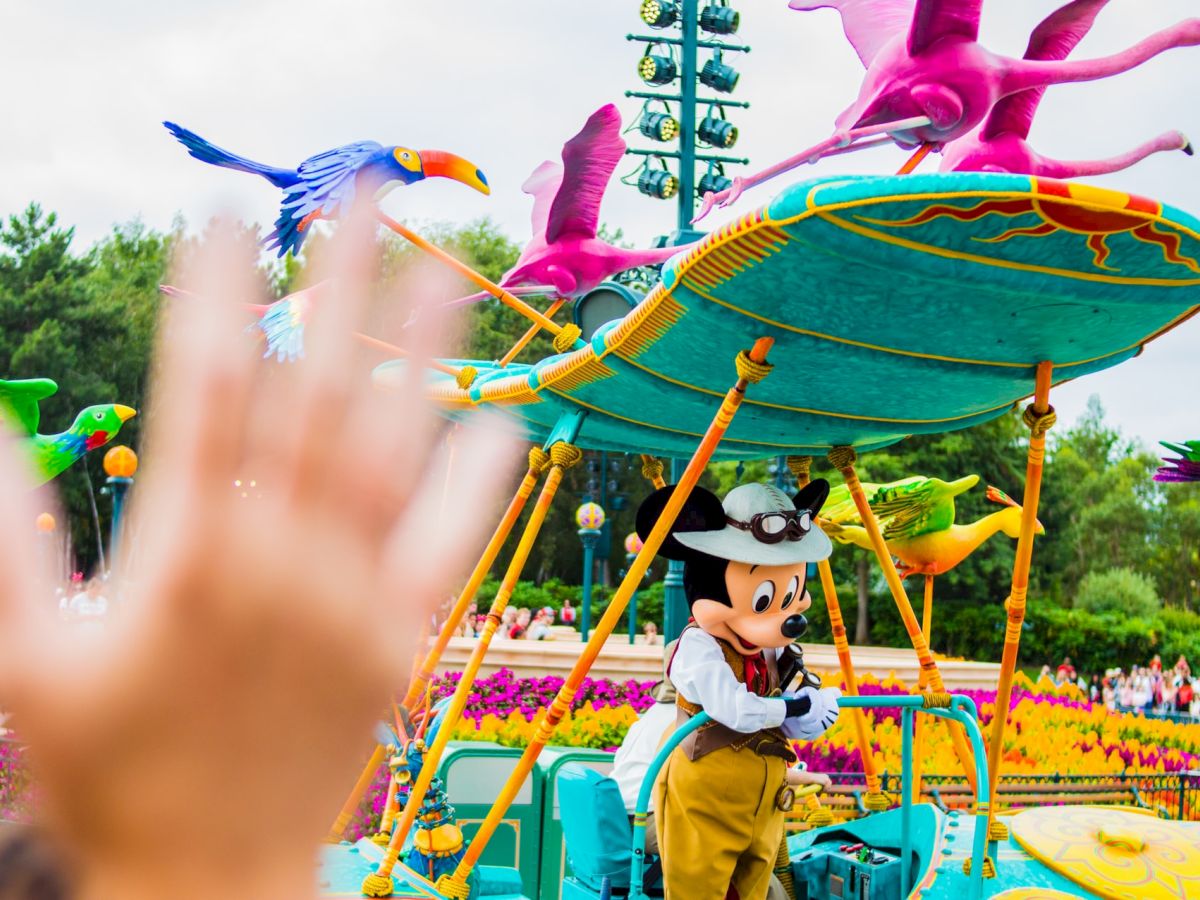 A costumed character is seen in a colorful amusement park setting with an outstretched hand in the foreground, waving.