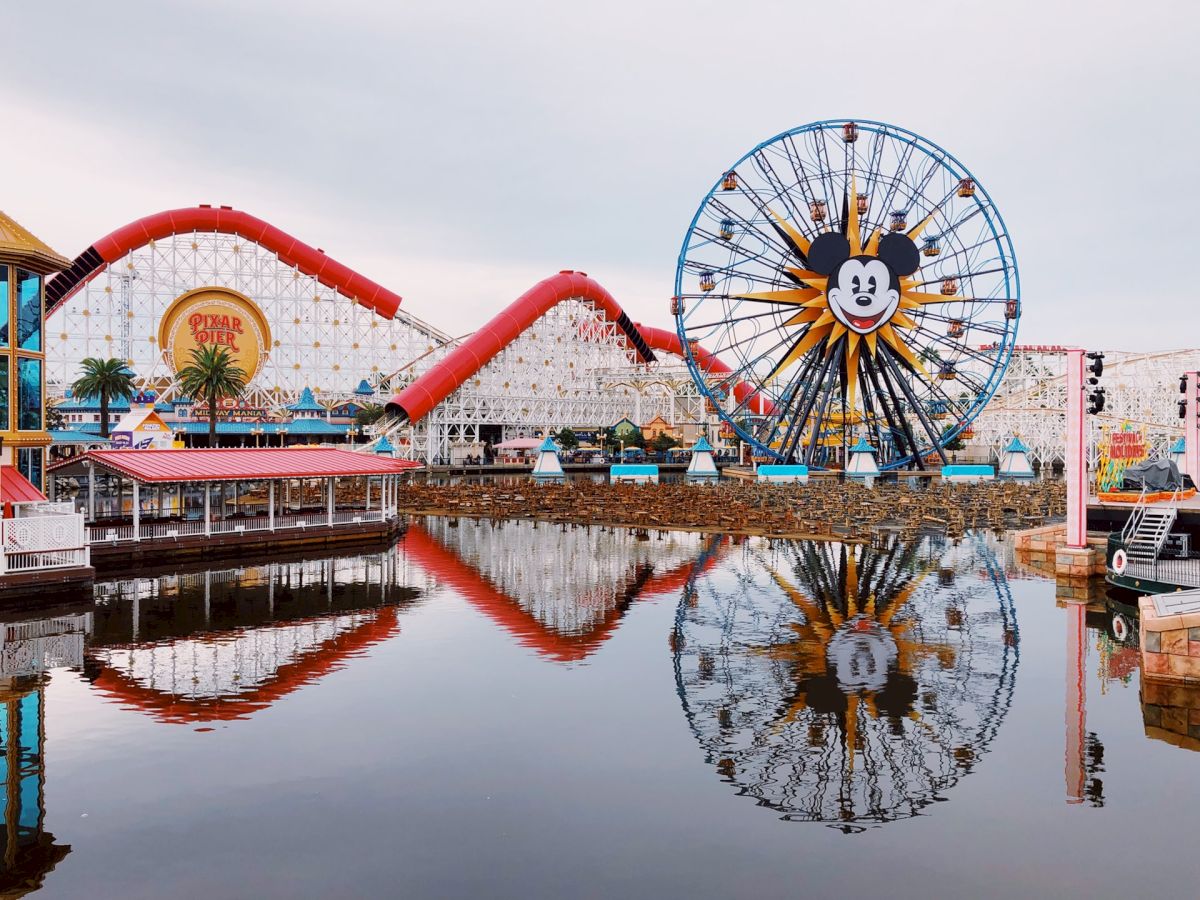 An amusement park with a Ferris wheel featuring a character's face, a roller coaster, and their reflections in calm water, creating a picturesque scene.