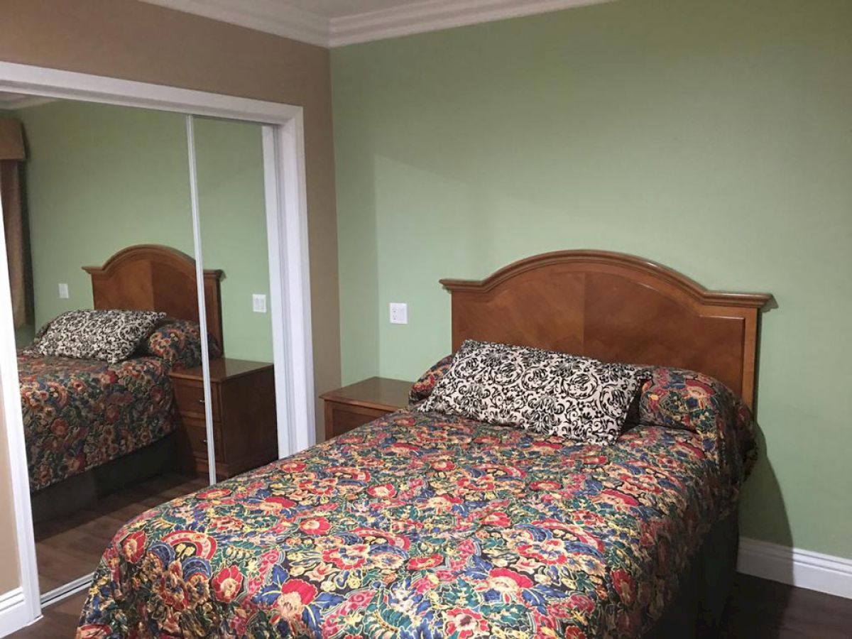 A small bedroom with a bed featuring a floral-patterned bedspread, a mirrored closet, green walls, and wooden furniture, including a nightstand.