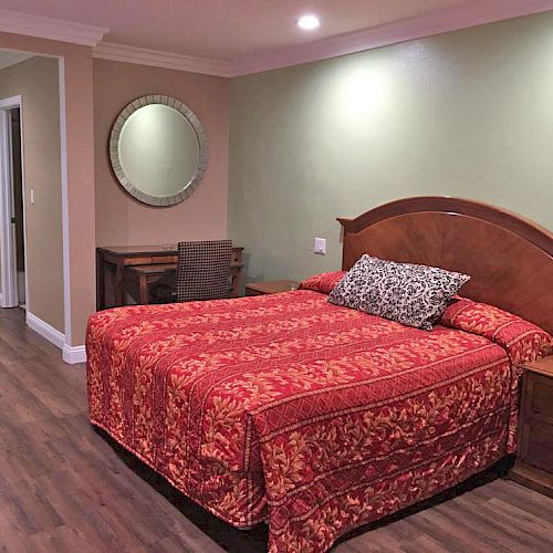 A cozy bedroom with a red-patterned bedspread on a wooden bed, a mirror, a desk, a chair, and wooden nightstands on hardwood flooring.