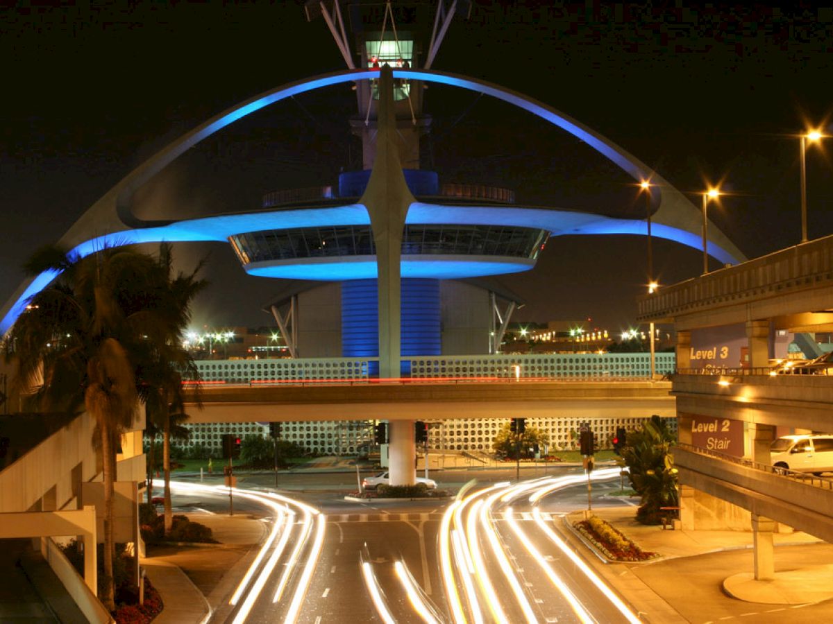 A futuristic structure illuminated at night with light trails from vehicles passing under and around it, surrounded by parking levels.