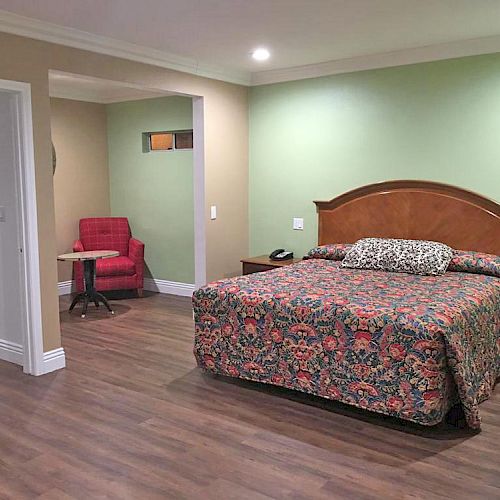 A hotel room with a floral-patterned bed, green walls, a red chair with a small table in the corner, wooden flooring, and a partially open door.