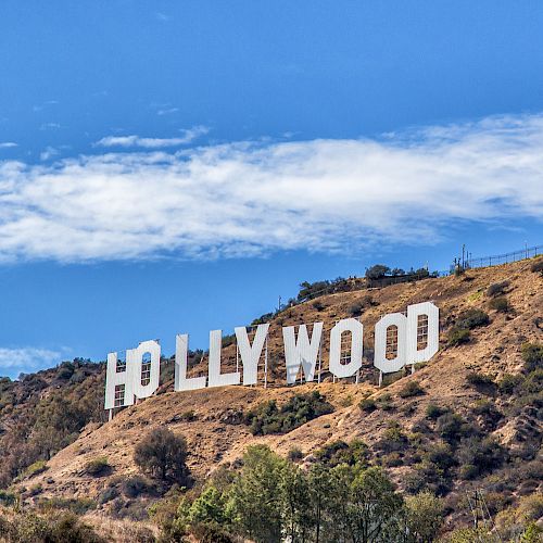 The image shows the iconic Hollywood sign in Los Angeles, California, set against a hilly background with a partly cloudy sky.