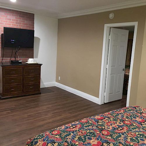 A bedroom with a patterned bedspread, a TV mounted on a brick wall, and a wooden dresser underneath. An open door is visible.