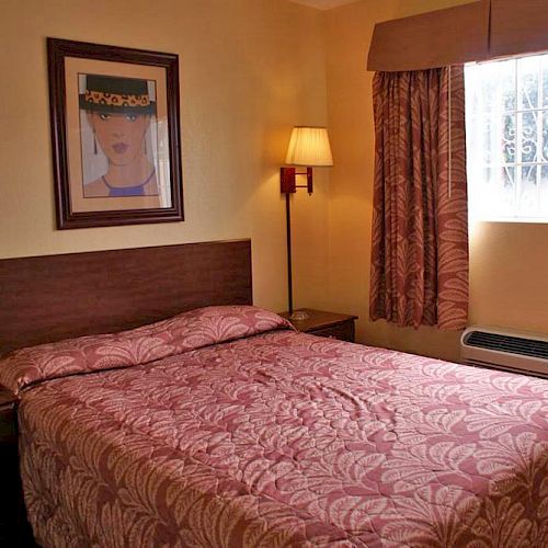 A cozy hotel room features a double bed, bedside tables, a lamp, framed art, patterned curtains, and a window with an air conditioner.