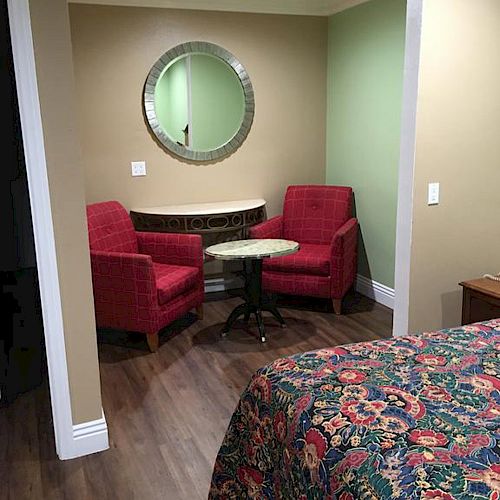 The image shows a cozy room with two red chairs, a small round table, a circular mirror, and part of a bed with a colorful bedspread.