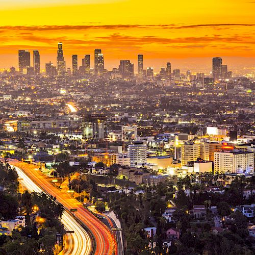 Cityscape at sunset featuring a bustling urban skyline and illuminated highways, with glowing orange and yellow skies creating a vibrant atmosphere.