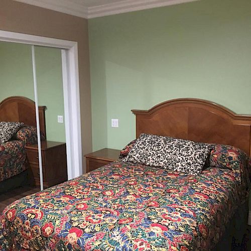 A cozy bedroom with a wooden headboard bed, colorful floral bedding, a patterned pillow, and a mirrored sliding closet door.