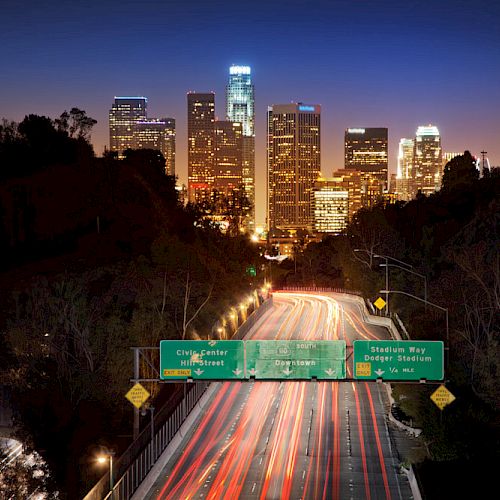 A cityscape at dusk with a brightly lit highway leading towards a downtown area filled with skyscrapers and illuminated street signs.