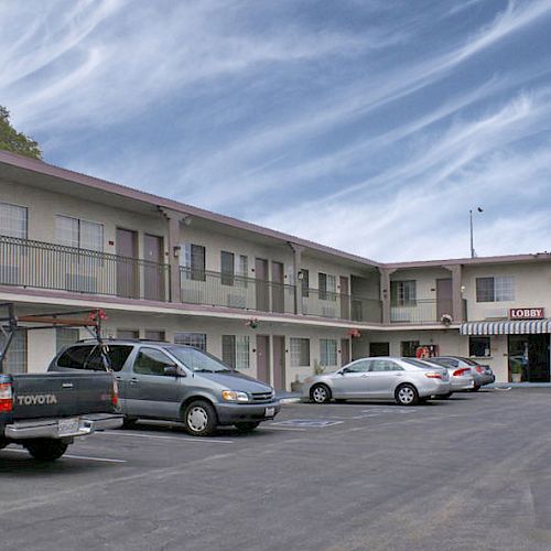 A two-story motel with several parked vehicles in front, including a pickup truck and a minivan, under a partly cloudy sky.