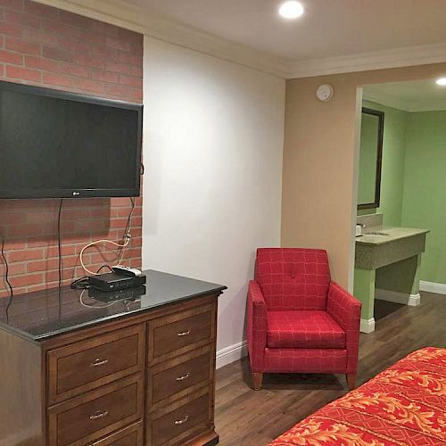 The image shows a hotel room with a wall-mounted TV, a wooden dresser, a red armchair, a bed with a red bedspread, and an adjacent sink area.