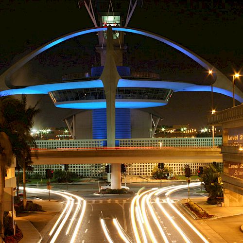 An illuminated futuristic building with arching structures at an airport with light trails from vehicles on the road below.