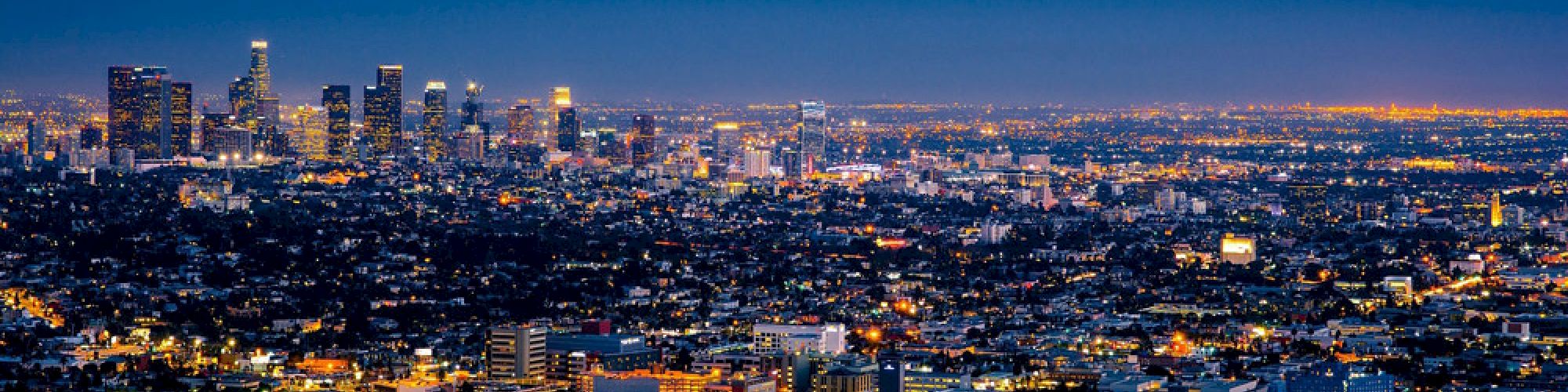 The image shows a sprawling cityscape at dusk, with numerous buildings and lights glowing under a dark blue sky, illustrating an urban setting.