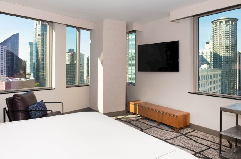 A modern hotel room with large windows showcasing a city view, a bed, TV, armchair, and wooden bench.