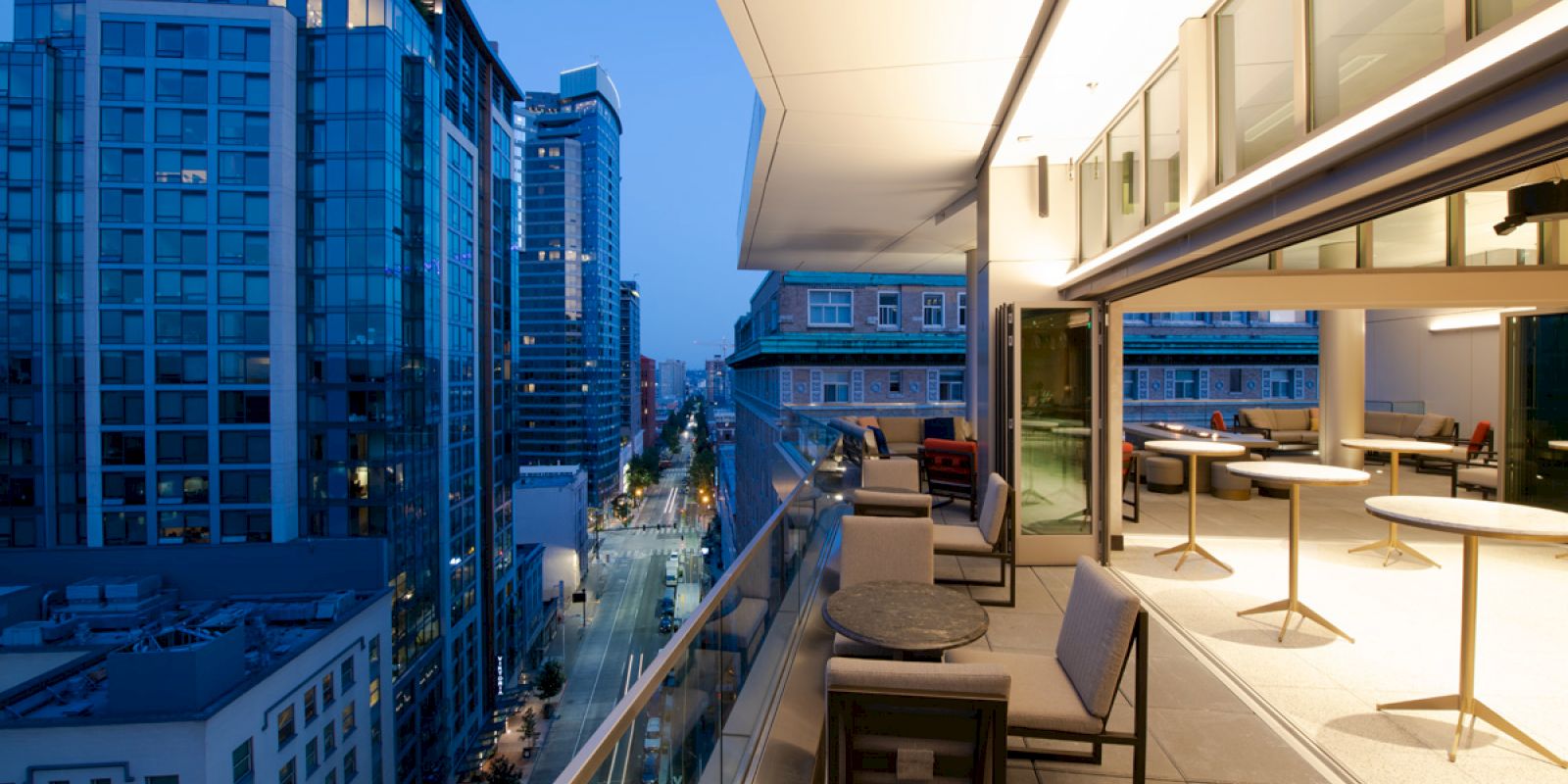 Modern cityscape view from a balcony at twilight, featuring buildings, a street, and outdoor seating with tables and chairs.