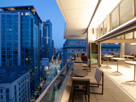 The image shows a modern cityscape at dusk from a high-rise balcony with seating, overlooking a street and neighboring buildings illuminated by lights.