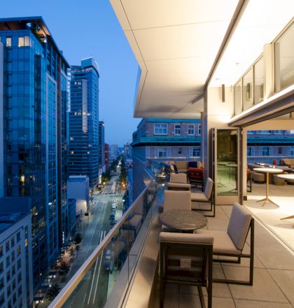 A modern cityscape at twilight with a view from a high-rise balcony featuring outdoor seating, looking down a lit street with tall buildings.