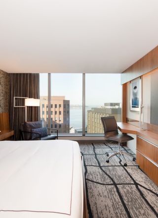 A modern hotel room with a large bed, flat-screen TV, desk, and seating area, featuring a view of city buildings through large windows.