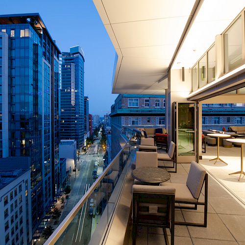 A modern cityscape at dusk with tall buildings and a spacious, well-lit balcony featuring outdoor seating and tables on the right side of the image.