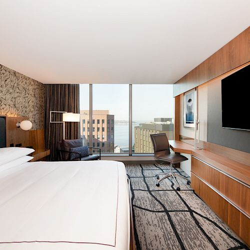 The image shows a modern hotel room with a large bed, a desk and chair, a wall-mounted TV, and a view of buildings and water through the window.