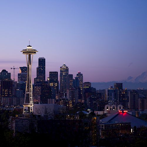 The image shows the Seattle skyline at dusk, featuring the Space Needle prominently and Mount Rainier in the background against a purplish sky.