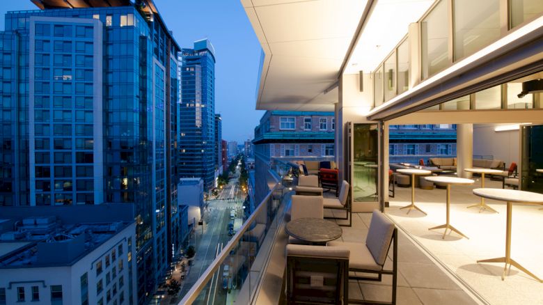 A cityscape at dusk featuring tall buildings, a street, and a modern balcony with tables and chairs on the right.