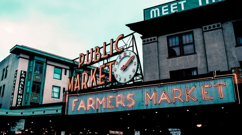 The image shows the iconic neon signs of Pike Place Market in Seattle, including "Public Market" and "Farmers Market," against an urban backdrop.