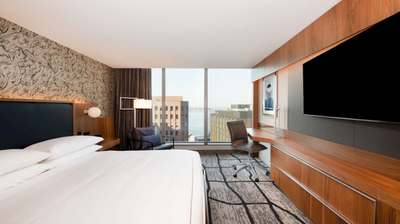 A neatly arranged hotel room with a large bed, desk, chair, TV, and a window offering a city view with buildings in the distance.